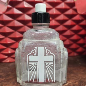 May include: Clear glass bottle with a white cross design and a black and white cap. The bottle has an Art Deco style with vertical lines. The cross is centered on the front of the bottle.