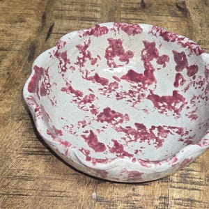 May include: A handmade ceramic bowl with a scalloped rim. The bowl is off-white with a speckled maroon pattern. The bowl is sitting on a wooden surface. This decorative bowl is ideal for serving food or as a decorative piece.
