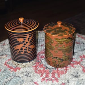 May include: Two ceramic jars with lids. One jar is black and orange with a spiral design on the lid and a pattern on the body. The other jar is orange with a brown and green mottled pattern. Both jars have orange knobs.