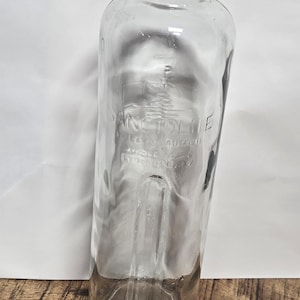 May include: Clear glass bottle with a wide base and a narrow neck. The bottle has embossed text that is difficult to read. The bottle is empty and appears to be vintage. The bottle is on a wooden surface.