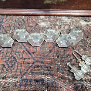 May include: A collection of clear, faceted glass cabinet knobs with metal hardware. The knobs are hexagonal and arranged on a patterned rug with red, brown, and blue hues. Several knobs are separated from their hardware.