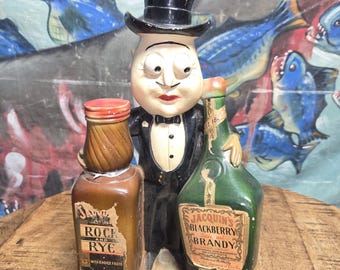 May include: A vintage Jacquin's Cordials figurine featuring a cartoon-like figure in a tuxedo holding two bottles. One bottle is labeled "Rock and Rye," and the other is "Blackberry Brandy." The figurine is on a wooden surface.