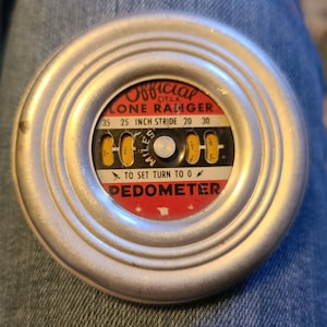 May include: A silver metal pedometer with a red and yellow dial. The dial reads "Official Lone Ranger 25 Inch Stride" and has a black arrow pointing to "To Set Turn To 0".
