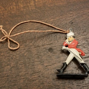 May include: A small, red and white figurine of a soldier with a sword, wearing a hat and holding a rifle. The figurine is attached to a brown string.
