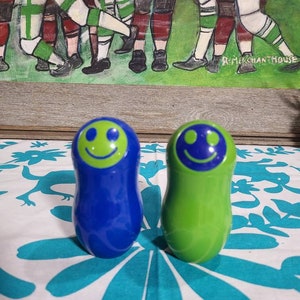 May include: Two nesting dolls, one blue and one green, with a smiling face on each. The dolls are on a white tablecloth with a turquoise floral pattern.