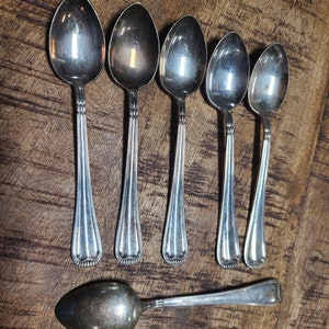 May include: Six silver spoons with ornate handles, laid out in a row on a wooden surface. A seventh spoon is lying horizontally below the others.