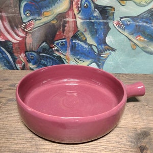 May include: A deep pink ceramic serving dish with a single handle. The dish is empty and has a smooth, glossy finish.