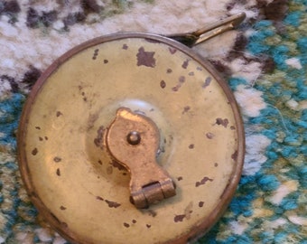 May include: A vintage metal tape measure with a brass handle and a worn, yellowed finish. The tape measure is closed and has a small, round hole in the center.