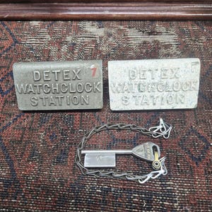 May include: Two rectangular metal DETEX Watchclock Station tags and a key with a chain. The tags are silver with black lettering and the number 7 on one. The key has a DETEX logo.