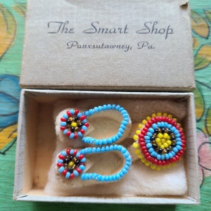 May include: A small box with a white interior and a beige lid. The lid has the text "The Smart Shop Punxsutawney, Pa." printed on it. Inside the box are two pairs of beaded moccasins and a beaded circle. The moccasins are blue and red with black and yellow beads. The circle is red, yellow, blue, and black.