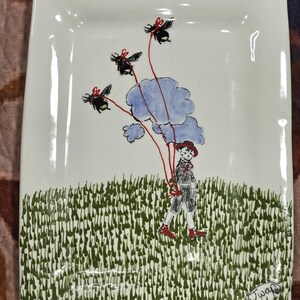 May include: A white ceramic rectangular serving plate with a hand-painted design of a child holding red balloons shaped like bumblebees. The child is wearing a red hat and blue pants. The plate has a green grass border and the signature "Juana" in the bottom right corner.