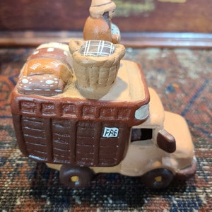 May include: A brown ceramic truck with a tan cab and a brown wooden bed. The truck is carrying a ceramic figure of a person wearing a hat and a basket of bread. The truck has the word "F&E" written on the side.