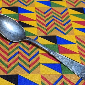 May include: A silver spoon with an ornate design on the handle. The spoon is lying on a yellow, red, blue, and black patterned fabric.