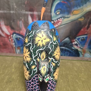 May include: Hand-painted wooden armadillo figurine with a black body, yellow and green accents, and a blue tail. The armadillo is standing on a green surface.