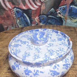 May include: A white and blue speckled ceramic serving dish with a lid. The dish is round with a small knob on top of the lid. The background features a painting of blue fish.