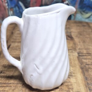 May include: A small, white ceramic pitcher with a handle and a spout. The pitcher has a textured, swirled design. The background is a wooden surface with a colorful painting.