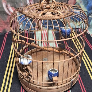 May include: A cylindrical bird cage constructed from thin brown bamboo slats with a circular wooden base. The cage features a metal hanging loop at the top, a curved wooden perch, and three small ceramic cups with blue and white floral patterns.