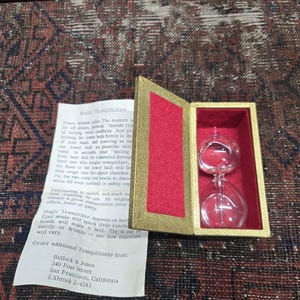 May include: A vintage Magic Tranquilizer in an open gold-colored box with a red interior. The hourglass-shaped device is made of clear glass. A paper with text about the product is visible.