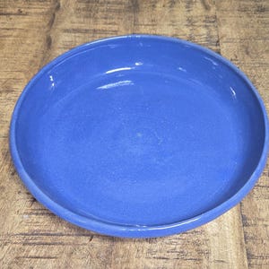 May include: A round, blue ceramic bowl with a wide rim. The bowl is centered on a wooden table with a natural wood grain finish. The bowl's interior has a smooth, glossy surface.