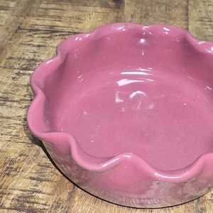 May include: A round, pink ceramic bowl with a scalloped edge. The bowl is a solid color and sits on a wooden surface. The bowl is likely used for serving or display.