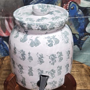 May include: A white ceramic water dispenser with a lid and spigot, decorated with a mottled blue-green pattern. It sits on a wooden stand, with a colorful painting in the background.