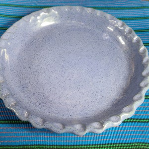 May include: A speckled, light blue ceramic pie plate with a scalloped edge. The plate has a slightly raised rim and a textured surface. The background is a blue and green striped fabric.