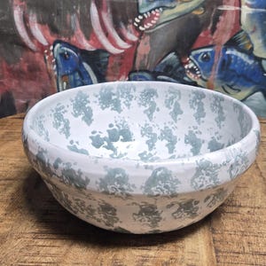 May include: A white ceramic bowl with a speckled blue-gray pattern. The bowl sits on a wooden surface, with a painted backdrop featuring blue fish with sharp teeth. The bowl is a decorative kitchen item.