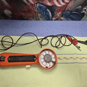May include: An orange rotary dial phone tester with black and red wires. The phone has a black dial with white numbers and letters. The tester is on a green and blue striped surface.