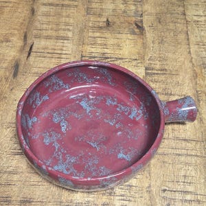 May include: A round, burgundy ceramic dish with a handle. The dish has a speckled blue and burgundy pattern. The background is a wooden surface with a painting of blue fish.