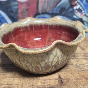 May include: A handmade ceramic bowl with a wavy rim. The interior is a glossy red, contrasting with the exterior's speckled tan and brown glaze. The bowl sits on a wooden surface.