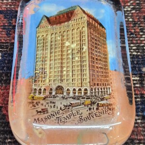 May include: Rectangular glass souvenir featuring a detailed illustration of the Masonic Temple building. The image shows the building's facade, with a blue sky background and the text "Masonic Temple Souvenir". The souvenir has a slightly curved shape.