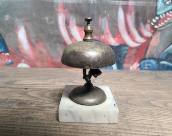 May include: A vintage brass bell with a marble base. The bell has a rounded top and a decorative stem with a small bell on each side. The bell is on a white marble base.