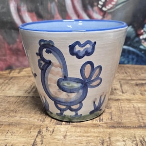 May include: A ceramic planter with a tapered shape and a white base. The exterior features a hand-painted blue outline of a rooster and a small cloud. The rim is painted with a solid blue band, and the interior is a light blue color.