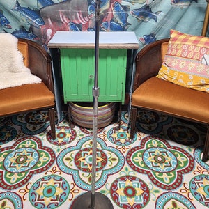 May include: A silver microphone on a stand with a black base. The microphone is in front of a green cabinet and two orange chairs with pillows. The background is a blue and white tapestry with fish.