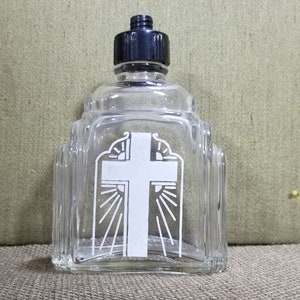 May include: Clear glass bottle with a black cap, featuring a white cross design. The bottle has a decorative, architectural shape with vertical ridges. The cross is centered on the front, with sun rays emanating from behind it.