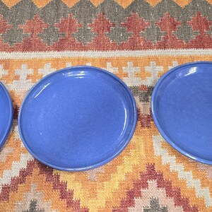 May include: Three round, blue ceramic plates are arranged on a colorful patterned rug. The plates have a slightly irregular shape and a glossy finish. The rug features a geometric design with shades of orange, red, and brown.