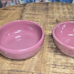 May include: Two small, round, pink ceramic bowls sit on a wooden surface. The bowls have a smooth, glossy finish and a simple, minimalist design. They are likely used for serving food or as decorative items.