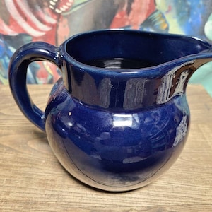 May include: A deep blue ceramic pitcher with a rounded body and a handle. The pitcher has a smooth, glossy finish.