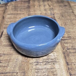 May include: A small, round, gray ceramic bowl with two handles. The bowl is centered on a wooden table with a natural wood grain finish. The bowl has a smooth, glossy interior.