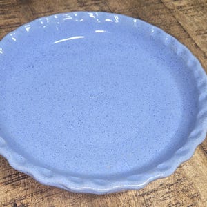 May include: A light blue ceramic pie plate with a speckled texture and a scalloped edge. The plate is round and sits on a wooden surface. The plate is approximately 10 inches in diameter.