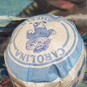 Op de afbeelding: Een vintage papieren hoed met de tekst "CAROLINA" in een blauw en wit cirkelvormig ontwerp. De hoed heeft een cartoon rammascotte in het midden, met een blauwe trui. De hoed staat op een mannequinhoofd.