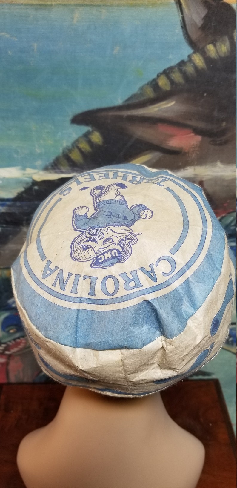 Op de afbeelding: Een vintage papieren hoed met de tekst "CAROLINA" in een blauw en wit cirkelvormig ontwerp. De hoed heeft een cartoon rammascotte in het midden, met een blauwe trui. De hoed staat op een mannequinhoofd.