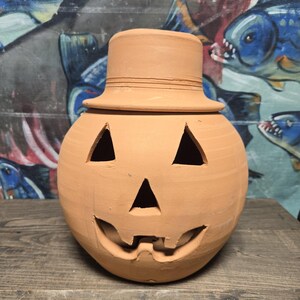 May include: A terracotta pumpkin-shaped lantern with a top hat lid. The lantern has triangular eye cutouts and a carved smiling mouth. The background features a colorful fish-themed mural.