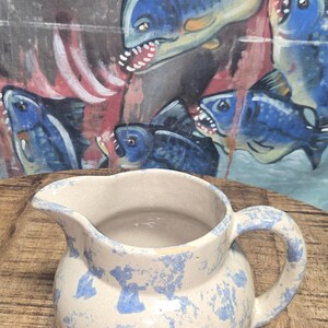 May include: A ceramic pitcher with a cream-colored base and blue splatter pattern. The pitcher has a rounded body, a curved handle, and a spout. The background features a painting of blue fish.
