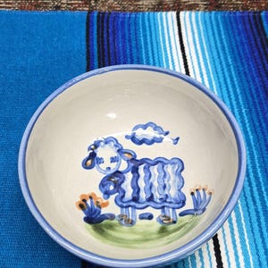 May include: A round ceramic bowl with a light cream-colored interior and a blue rim. The center of the bowl features a hand-painted blue sheep standing on a patch of green grass with small orange and blue floral accents and a blue cloud above.