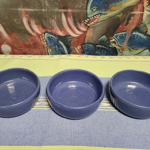 May include: Three blue ceramic bowls with a white glaze on a yellow and blue patterned surface. The bowls are round and have a slight lip.