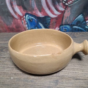 May include: A beige ceramic serving dish with a single handle. The dish is oval shaped and has a smooth, slightly textured surface.