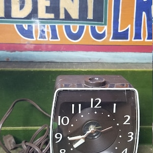 May include: A brown vintage electric alarm clock with a black face and white numbers. The clock has a cord and plug. The clock is set to 11:55.