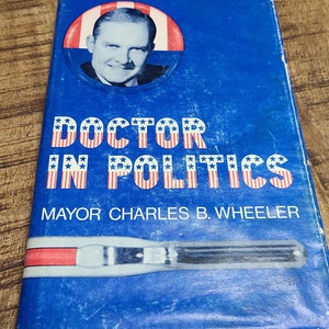 May include: A blue book with a red, white, and blue American flag design. The book has the title "Doctor in Politics" and the author's name "Mayor Charles B. Wheeler". A photo of a man in a button-up shirt is in the upper left corner.