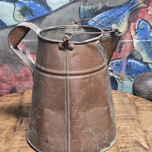 May include: A vintage copper pitcher with a handle and spout. The pitcher has a cylindrical body with a wide base and a narrow top. The copper has a weathered patina, and the handle is attached with rivets. The background features a colorful fish painting.